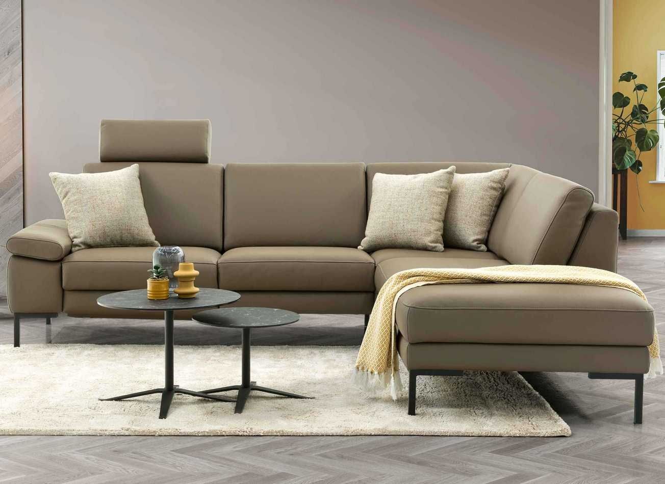 Ecksofa aus Leder mit verstellbarer Kopfstütze, zwei Kissen und passendem Hocker auf hellem Teppich vor grauer Wand