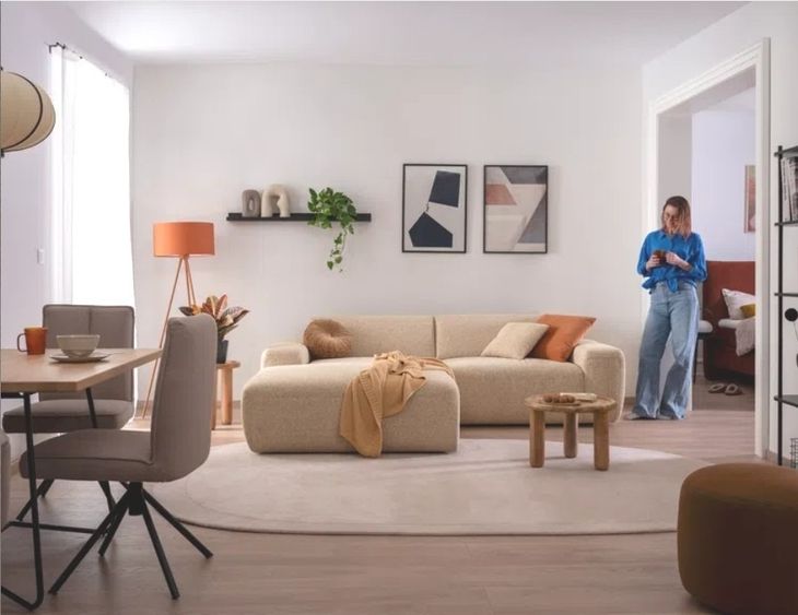Wohnzimmer mit hellem Sofa, kleinem Holztisch, Stehlampe, Wandregal mit Pflanze und zwei abstrakten Bildern, daneben steht eine Frau in blauer Bluse und Jeans